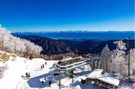 雪山の風景写真です。雪で覆われた山と木々が見えます。遠くには山脈が連なっています。手前にはリフトの乗り場とスキーを楽しむ人々が写っています。空は青く晴れています。