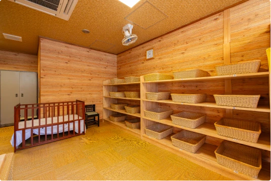 木の温もりを感じる脱衣所。木製の壁と床、籐製の脱衣かごが並ぶ棚、ベビーベッド、小さな椅子、時計、換気扇が写る。