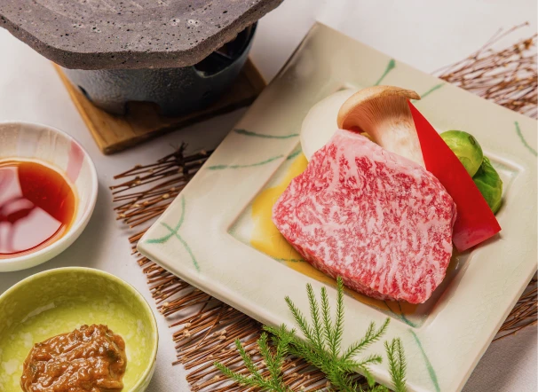 会席料理の一つ。中央に陶板焼きの皿、牛の赤身肉と野菜、椎茸、ネギが並ぶ。左にタレの入った小鉢二つ、右に松の葉の飾り。