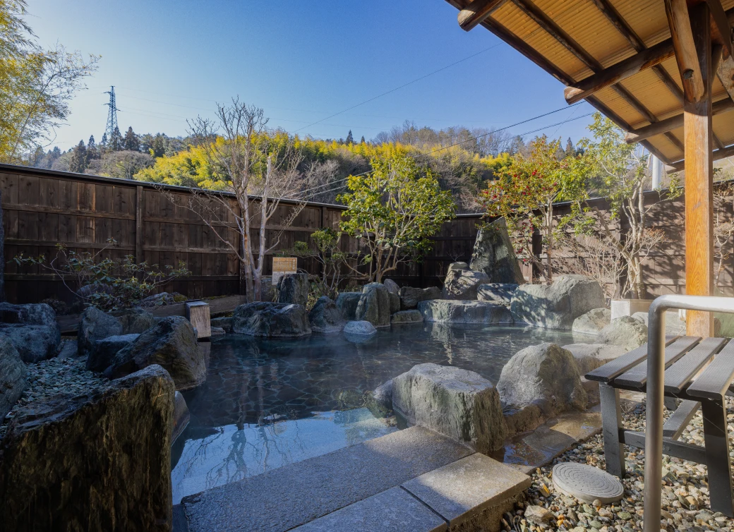 木々に囲まれた露天風呂。大小の岩が点在する湯船から湯気が立ち上り、穏やかな陽光が水面を照らしている。