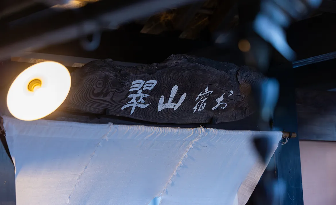 木製の看板に「お宿 山翠」と書かれている。看板の下には白い暖簾がかかっている。