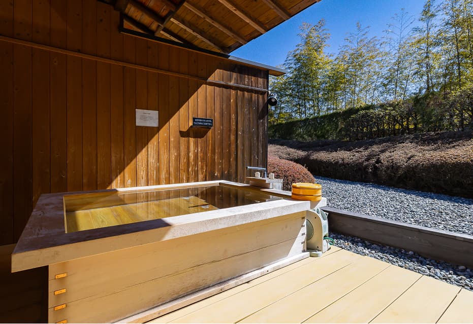木材をふんだんに使用した、温かみのある露天風呂。湯船には温泉が張られ、湯面が光を反射しています。露天風呂の床は木製のデッキで、湯船の奥には緑豊かな庭園が広がっています。